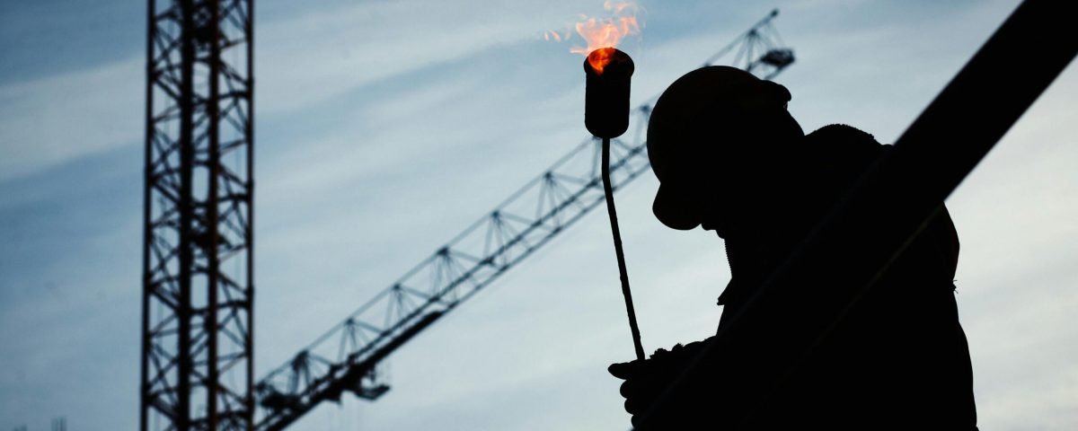 Silhouette of a construction worker using a blowtorch at a building site against a crane-filled skyline.